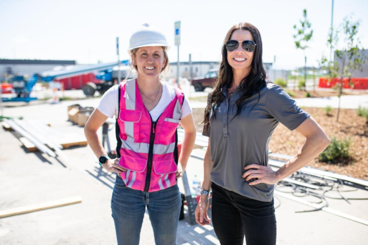 A woman wearing shades standing next to a woman in a pink high vis vest and white helmet