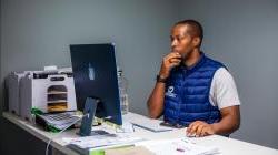 Man working at a desk with an iMac, wearing a blue vest, surrounded by papers and office supplies.