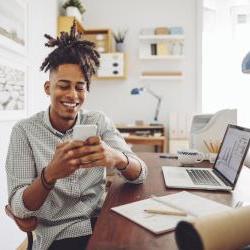 young black man in button down sitting at computer and laughing at phone