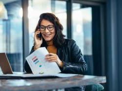 Woman sitting at table with laptop reviewing finances as she talks on her cellphone